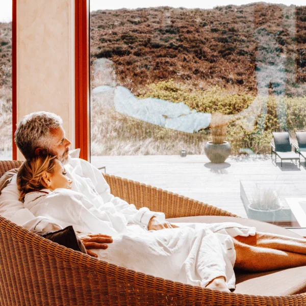 Ein Paar entspannt sich in einem hellen und gemütlichen Wellnessbereich, eingehüllt in weiße Bademäntel und in einem großen Rattansessel sitzend. Sie lehnen sich zurück und genießen die ruhige Aussicht durch eine große Fensterfront, die den Blick auf eine friedliche Dünenlandschaft mit einigen Sonnenliegen im Freien freigibt.