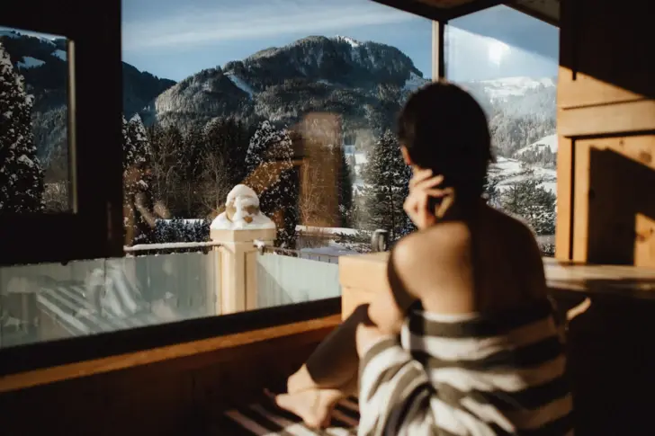 Eine Frau sitzt in ein Handtuch gewickelt in einer hölzernen Sauna mit Glasfront auf die schneebedeckten Berge und lässt sich die Sonne ins Gesicht scheinen. 