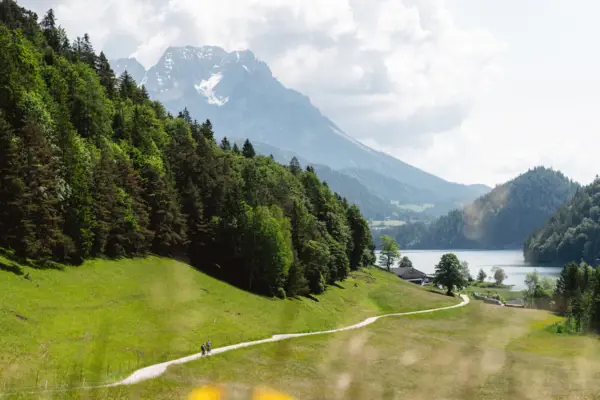 Pfad führt durch eine Wiese zu einem See, umgeben von Bergen und Himmel mit Wolken.