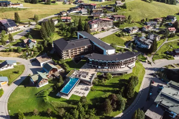 Großes Gebäude umgeben von Grasflächen und Häusern im alpinen Stil, aufgenommen aus der Vogelperspektive.