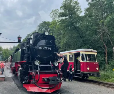 Dampflokomotive der Harzer Schmalspurbahn am Bahnhof, daneben ein historischer Triebwagen. Menschen steigen ein und aus, während die Lokomotive dampft und von Mitarbeitern betreut wird.