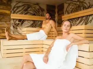 Ein Mann und eine Frau entspannen, in weiße Handtücher gewickelt in einer Holzsauna. An der Wand befindet sich Dekoration aus Treibholz. 