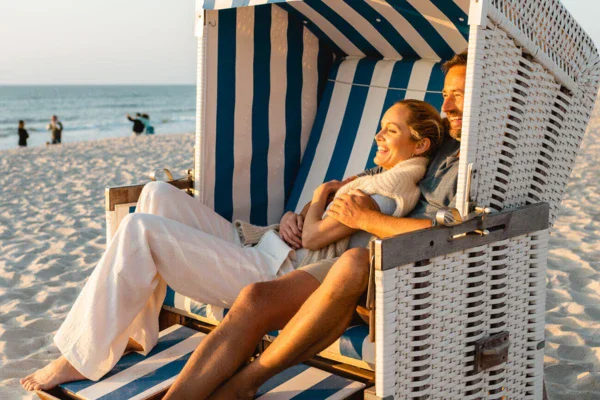 Ein Mann und eine Frau sitzen lächelnd, Arm in Arm, bei Sonnenuntergang, in einem weiß blau gestreiften Strandkorb. Die Sonne scheint warm und golden auf ihre Haut.