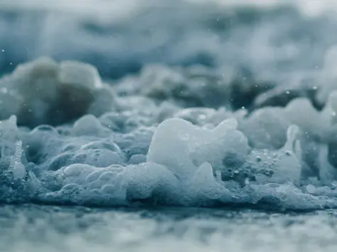 Detailaufnahme von sprudelndem Wasser, das im Licht schimmert, mit einem Fokus auf die einzelnen Wasserblasen und den entstehenden Schaum auf der Wasseroberfläche.