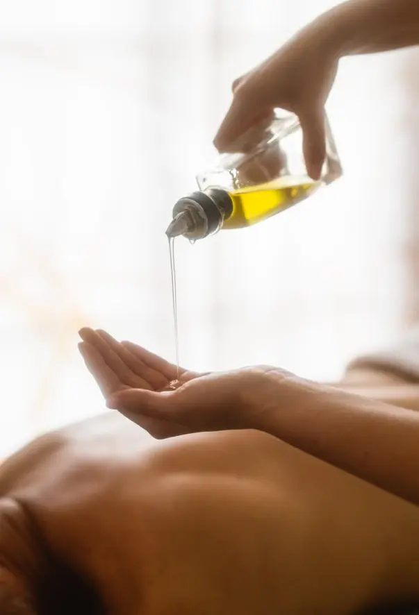 Massageöl wird aus einer Flasche auf eine wartende Hand gegossen. Der Rücken einer Person ist im Hintergrund zu sehen, die für eine Massage auf einer Liege liegt. 