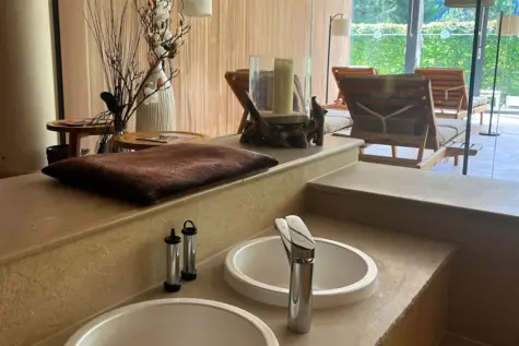 Eleganter Spa-Bereich mit zwei weißen Waschbecken auf einer Steinplatte, stilvollen Deko-Elementen wie Trockenblumen und Kerzen sowie gemütlichen Holzliegen im Hintergrund mit Blick auf eine sonnige Terrasse und grüne Hecke.