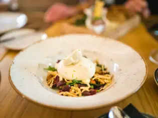 Ein elegantes Gericht mit Burrata und Gemüse wird auf einem stilvoll angerichteten Teller serviert, während eine Person mit einer Gabel das Essen genießt. Im Hintergrund stehen weitere Speisen, Rotwein und Gläser auf einem Holztisch.