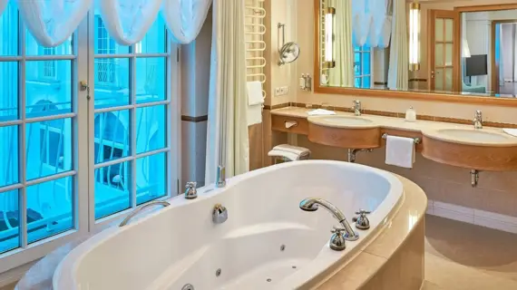 Luxurious bathroom in a suite with a large bathtub and a large window in front of it with translucent white curtains that let in natural light. The room is equipped with two washbasins on a marble counter, an illuminated wall mirror and high-quality fittings. A combination of warm beige and brown tones and the soft textiles create an elegant and relaxing ambience.