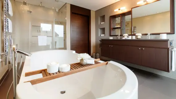Ein großes, modernes Badezimmer mit einer Badewanne im Vordergrund. Im Hintergrund ist eine verglaste Dusche sowie ein langer dunkler Waschtisch mit zwei eingelassenen Waschbecken zu sehen.