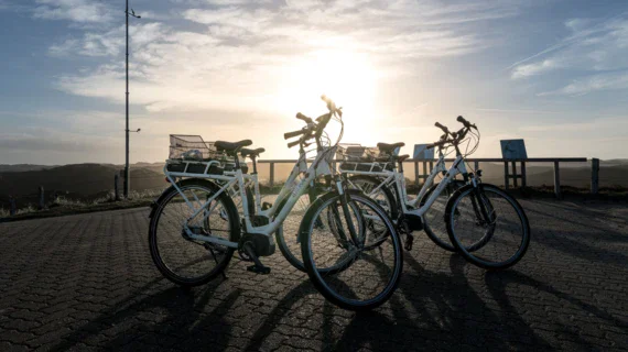 Drei weiße Fahrräder stehen geparkt auf einer gepflasterten Fläche mit Blick auf eine hügelige Landschaft im Gegenlicht der untergehenden Sonne. Eine friedliche Szene mit klarem Himmel und sanften Konturen der Hügel im Hintergrund.