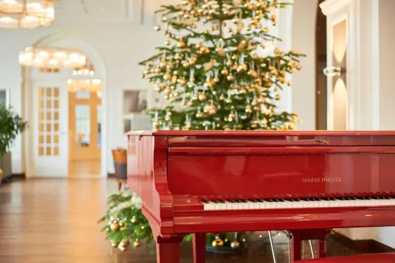 Ein roter Flügel der Marke August Förster steht festlich geschmückt vor einem goldverzierten Weihnachtsbaum in einem elegant beleuchteten Raum.
