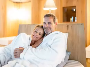  Ein lächelndes Paar in weißen Bademänteln kuschelt gemütlich auf einer Bank in einem ruhigen, hellen Ruheraum mit Holzinterieur.