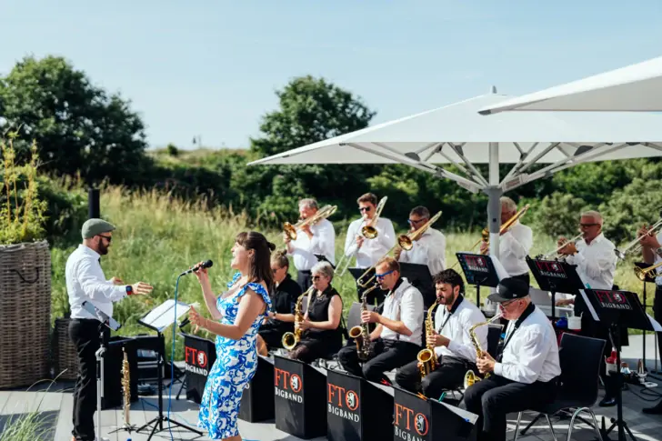 Ein Musiker springt mit ausgebreiteten Armen in die Luft, während das Orchester lachend im Hintergrund auf einer sonnigen Hotelterrasse steht.