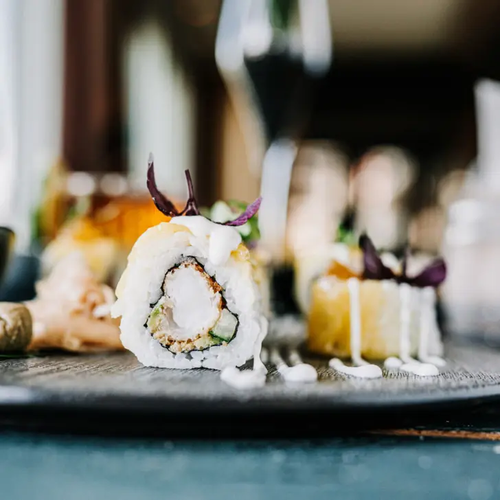 Im Fokus steht eine mit Garnele und Gurke gefüllt Sushi Rolle. Diese ist aufgeschnitten und mit anderem Sushi auf einem dunklen Teller angerichtet. 