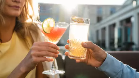 Zwei Personen stoßen bei Sonnenuntergang auf einer Terrasse mit Getränken an. Im Fokus steht eine Hand, die einen Cocktail mit Limettenscheibe in einem Martini-Glas hält, und eine andere Hand, die ein Highball-Glas mit einem klaren, sprudelnden Getränk und Orangenscheibe hält. Die warme Abendsonne beleuchtet die Szene, was eine entspannte und gesellige Stimmung vermittelt.