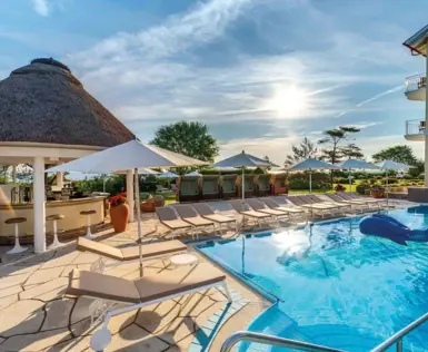 Entspannender Außenbereich mit Pool, Liegestühlen und Sonnenschirmen, begleitet von einer gemütlichen Poolbar mit Reetdach. Die Abendsonne spiegelt sich im Wasser, während der Blick auf eine gepflegte Gartenlandschaft und den weiten Himmel fällt.