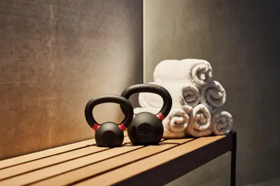  The image shows a section of a fitness room with a focus on two black kettlebells with red markings placed on a wooden bench. A stacked pyramid of six neatly rolled white towels can be seen behind the kettlebells.