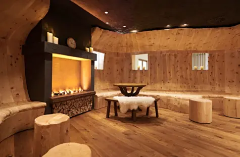 Ein gemütlicher Raum mit holzverkleideten Wänden und Boden, ausgestattet mit einem Kamin, Kerzen, einem rustikalen Holztisch, Hockern und einer Bank mit einem flauschigen Lammfell. Das Ambiente ist warm und einladend mit indirekter Beleuchtung.