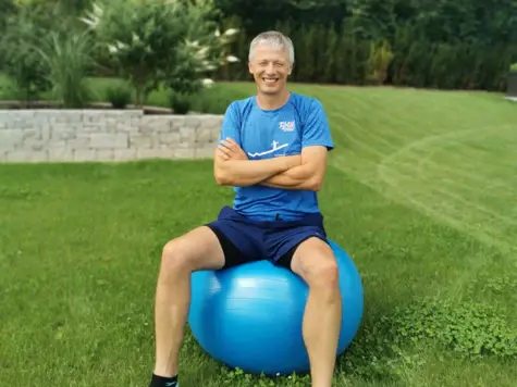 Ein Mann sitzt draußen auf einem großen blauen Gymnastikball auf einer Wiese.