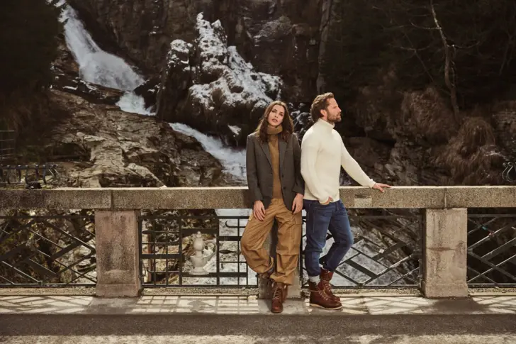 Zwei stilvoll gekleidete Personen stehen auf einer Brücke vor dem beeindruckenden Gasteiner Wasserfall in Bad Gastein und genießen den winterlichen Ausblick in alpiner Kulisse.