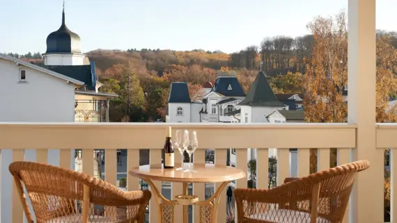 A peaceful balcony view of picturesque white buildings with distinctive black roofs and a church steeple, nestled in a sea of autumn-colored trees. In the foreground is a rattan table with two matching chairs, perfectly positioned to enjoy the view. On the table is an open bottle of wine with two empty glasses, suggesting a relaxing atmosphere. The golden fall light enhances the warm and inviting feeling of this scenario.