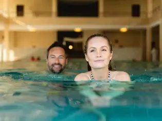 Eine Frau und ein Mann schwimmen lächelnd in einem Innenpool.