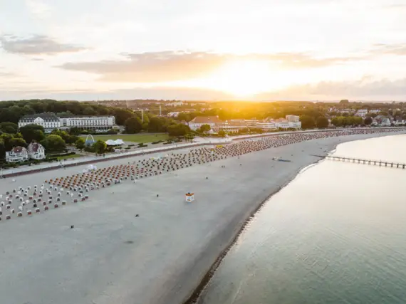 Eine Luftansicht von einem langen Strand am Wasser und dahinterliegende Gebäude im Morgentau bei Sonnenaufgang.