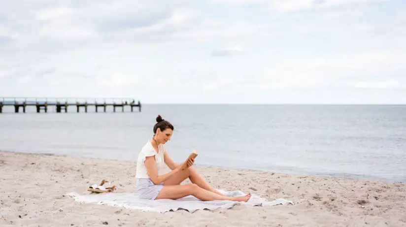 Frau sitzt auf einem Handtuch am Strand und liest ein Buch.