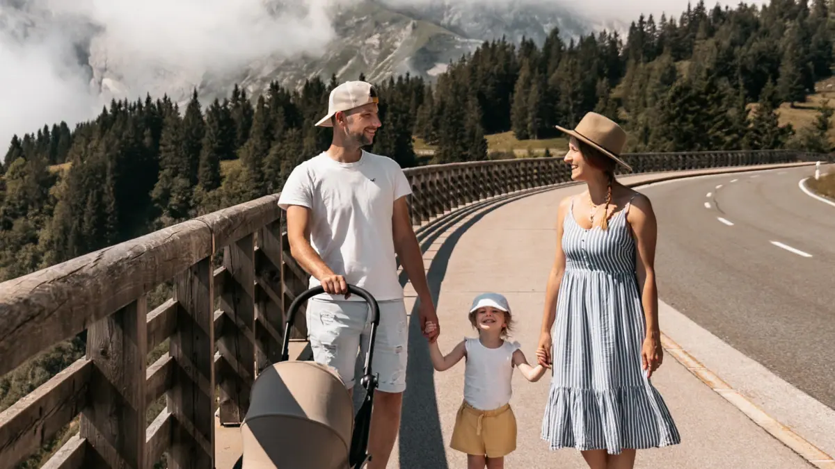 Eine Familie geht mit einem Kinderwagen über eine Brücke, im Hintergrund ist ein Berg zu sehen.