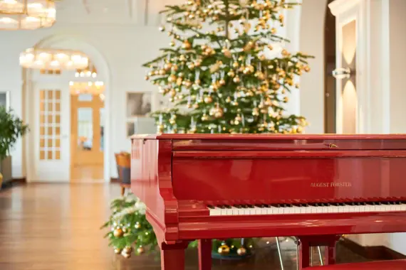 Ein festlich geschmückter Raum mit einem roten August Förster-Klavier im Vordergrund und einem strahlenden Weihnachtsbaum voller goldener Kugeln im Hintergrund