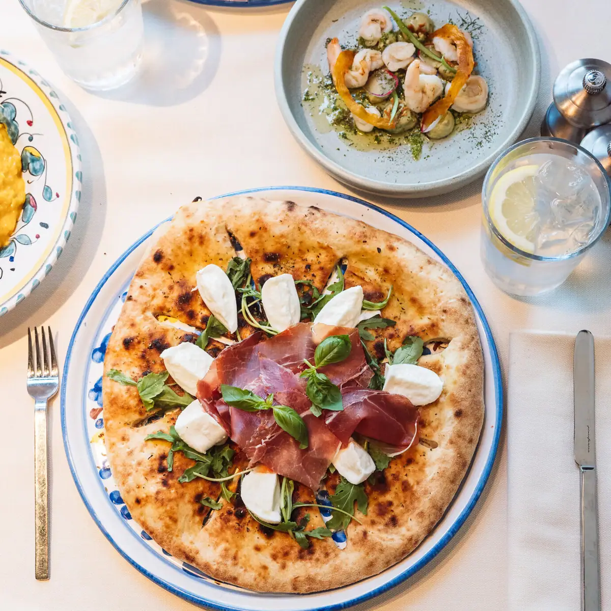 Ein Teller mit italienischer Pizza auf einem Tisch. Im Hintergrund sieht man ein Wasserglas sowie Scampi auf einem tiefen Teller.