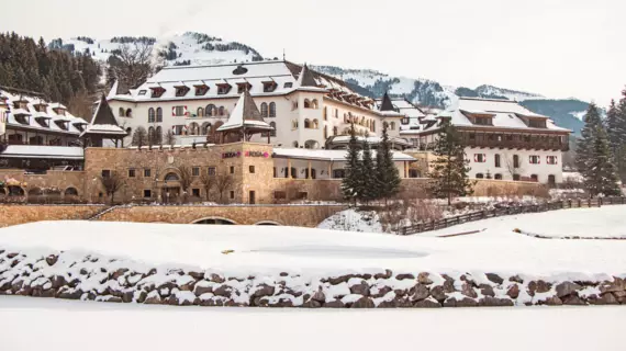 Das Bild zeigt das große, traditionelle Gebäude des A-ROSA Kitzbühel mit einem alpinen Architekturstil, das von Schnee bedeckte Berge im Hintergrund überragt. Das Hotelgebäude ist weiß verputzt mit dunklen Holzverkleidungen und mehreren Türmchen, die ihm ein schlossähnliches Aussehen verleihen. Es scheint ein Wintertag zu sein, da der Boden vor dem Gebäude mit Schnee bedeckt ist und die Bäume um das Gebäude herum kahl sind. Auf der Fassade des Gebäudes ist ein Schild mit der Aufschrift "A-ROSA" zu sehen.