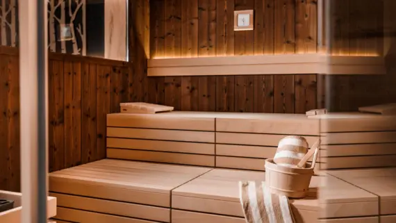 Eine Sauna mit zwei Bänken im gemütlichen Licht. Auf der unteren Saunabank steht ein Holzkübel für Aufgüsse und zwei gestreifte Saunahandtücher.