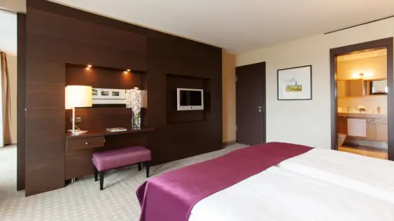 A hotel room with a large bed on which a purple bedspread lies. The view falls on a wall covered with dark wooden panels. There is a desk made of the same material as the wall with a lamp and flowers on it. The door to the room and access to the bathroom can be seen on the left.
