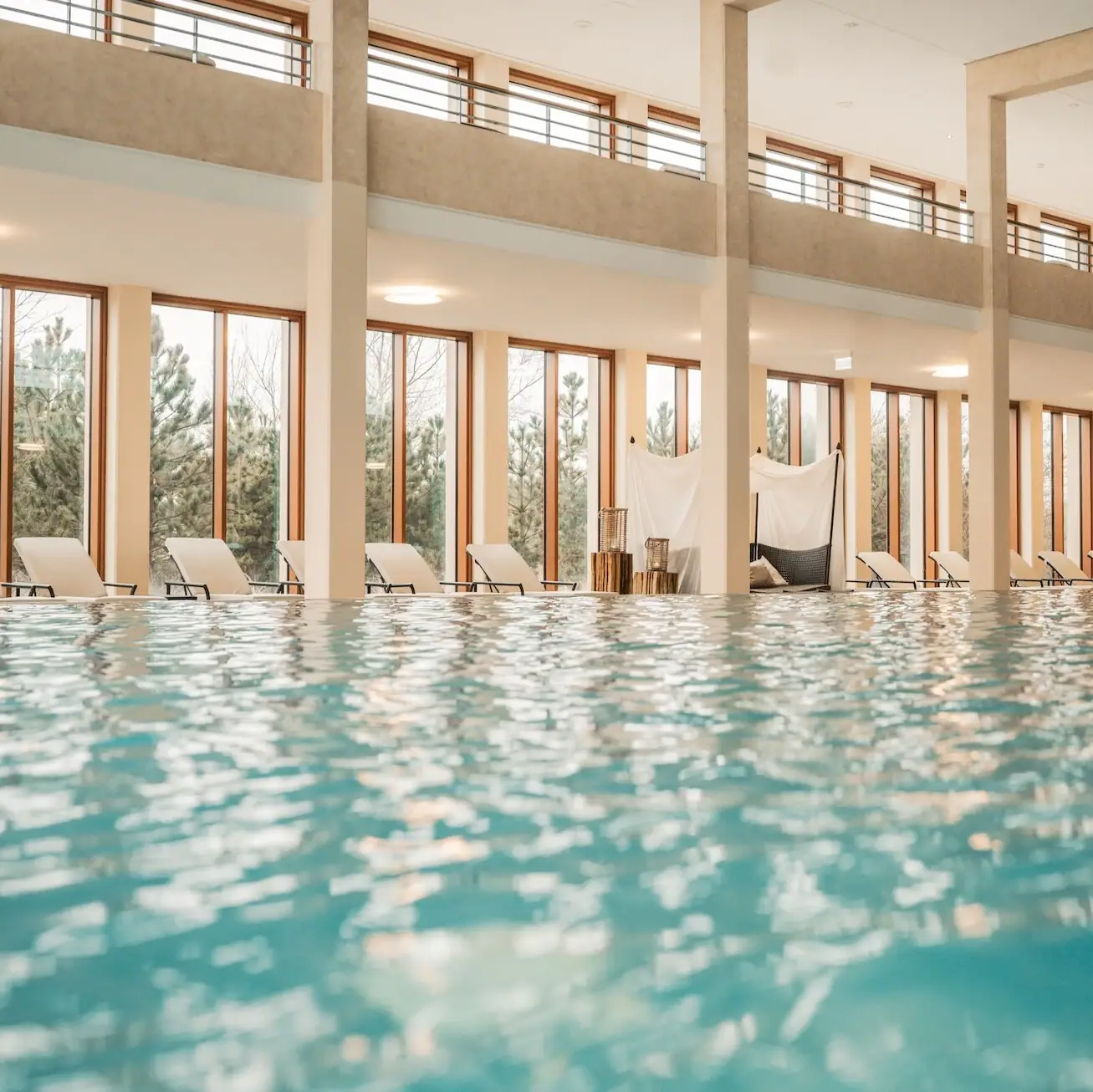 Ein Schwimmbecken mit Liegestühlen am Rand und Ausblick auf die Dünenlandschaft durch die Fenster im Hintergrund..