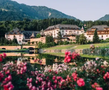  Ein elegantes Resortgebäude mit mehreren Stockwerken und einem Schindeldach, eingebettet in eine grüne Landschaft mit einem Hintergrund aus bewaldeten Hügeln. Im Vordergrund spiegelt sich das Gebäude in einem ruhigen Teich, umrahmt von blühenden Rosen.