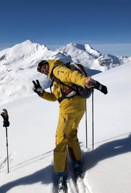Ein Mann in einem gelben Skianzug steht auf einem verschneiten Berg mit Skiausrüstung.