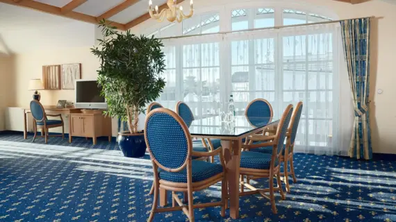 A spacious and bright living room flooded with natural light from the large windows. The room is tastefully furnished with a large round wooden dining table and matching chairs with blue upholstery. The deep blue carpet with yellow floral patterns enhances the maritime flair of the room. On the left of the picture is a work area with a large desk and modern furnishings. Green plants provide freshness and a connection to nature, while the classic chandelier and fine curtains emphasize the luxurious character of the room. The harmonious combination of traditional elements and modern comfort conveys an atmosphere of exclusive serenity.