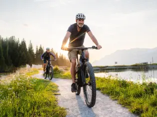 Zwei Radfahrer fahren auf einem schmalen Pfad entlang eines Bergsees in den Kitzbüheler Alpen. Die Sonne scheint durch die Bäume.