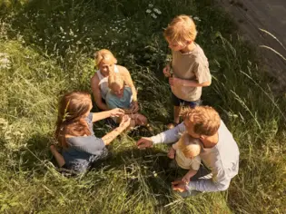 Ein Erwachsener trägt ein Kind auf den Schultern, beide mit Hüten bekleidet, während sie durch eine malerische Berglandschaft mit Wiesen und Wald spazieren.