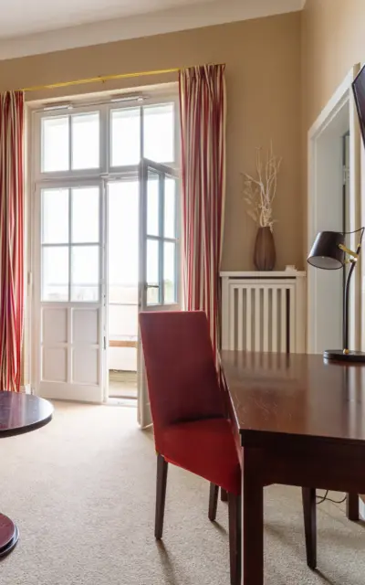  Ein heller und einladender Wohnzimmerbereich mit einem großen Fenster, das viel Tageslicht hereinlässt. Das Interieur ist in warmen Beige- und Erdtönen gehalten, mit einer großen modernen Couch und roten Akzentkissen, die einen Farbtupfer bieten. Ein dunkler Holztisch und ein dazu passender Stuhl stehen im Raum, darauf eine stilvolle Lampe und eine Vase mit dekorativen Zweigen. Ein großes, abstraktes Wandbild bringt zusätzliche Farbe und Textur in den Raum. Die langen, gestreiften Vorhänge rahmen das Fenster ein und verleihen dem Raum eine gemütliche Atmosphäre.