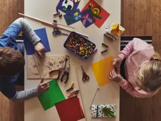 Zwei Kinder sitzen an einem Tisch und basteln mit buntem Papier, Holzstäben, Perlen und anderen Bastelmaterialien, während sie kreativ und konzentriert arbeiten.