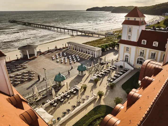 Eine Luftansicht des Kurhauses Binz mit seinen weißen Fassaden und roten Dächern. Im Innenhof stehen Stühle und Schirme, die Sonne taucht den Tag in eine frische Morgenluft, während das Meer im Hintergrund leichte Wellen schlägt. 