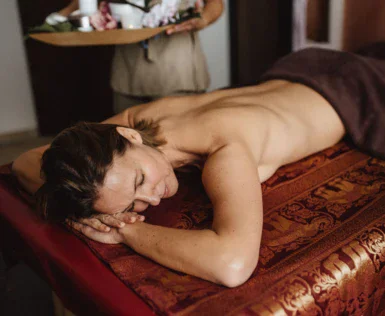 Eine entspannte Person liegt auf dem Bauch auf einer Massageliege, bedeckt mit einem Handtuch, während im Hintergrund eine Mitarbeiterin mit einem Tablett voller Wellnessprodukte steht.