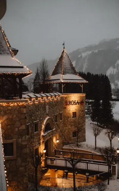 Ein gemütliches Hotel aus Naturstein mit beleuchteten Türmen und weihnachtlicher Beleuchtung, umgeben von verschneiten Bäumen und Bergen in einer winterlichen Landschaft.
