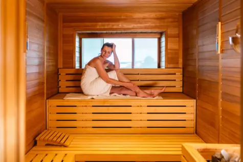 Eine Person, eingehüllt in ein Handtuch, sitzt entspannt und lächelnd in einer holzverkleideten Sauna, die durch ein Fenster einen Ausblick auf die Umgebung bietet.