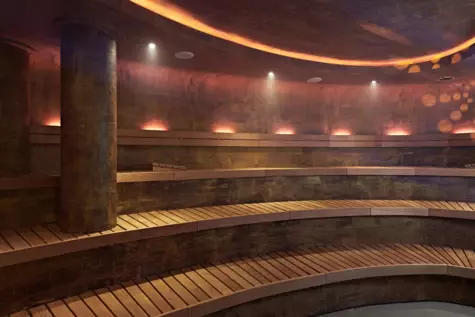 Ein stimmungsvoller Saunaraum mit kreisförmigen Sitzbänken aus Holz, strukturierten dunklen Wänden und einer beleuchteten Decke, die eine warme und ruhige Atmosphäre erzeugt.
