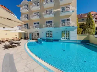 Loungers are lined up around the outdoor pool at the A-ROSA Binz and the sun shines warmly on the water. The adjacent hotel building stands in the background.