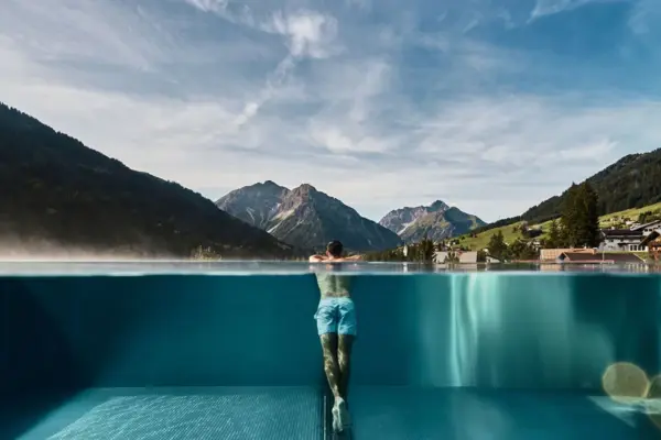 Mann lehnt am Rand eines Infinity-Pools mit Blick auf einen Ort inmitten einer grünen Berglandschaft im Hintergrund.