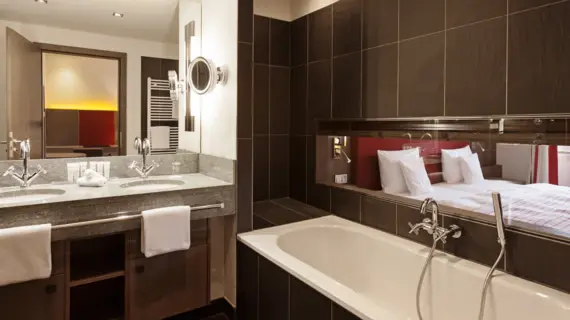 Ein Badezimmer mit dunklen Fließen, einer Badewanne und einem Waschtisch mit zwei Waschbecken. Durch ein schmales Fenster über der Badewanne ist ein Bett im Nebenraum zu sehen.